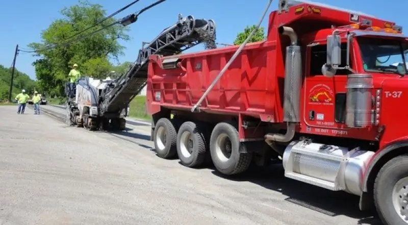 A red Shallenberger tri-axle dump truck, unit T-37, beside a milling machine with crew in hi-vis