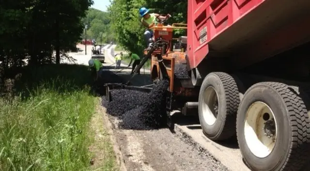 A dump truck feeding a paver on a narrow road, hot asphalt in the hopper