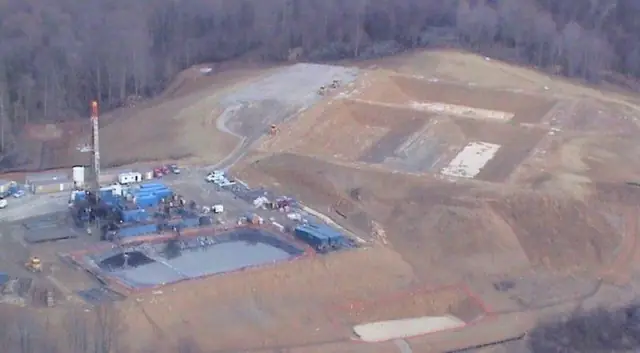 Aerial of a well site with a rig mast, graded pads and a lined pond in the woods