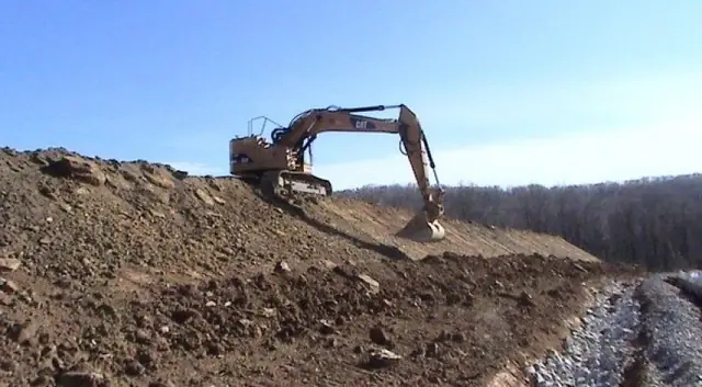 An excavator on a cut slope above a riprap-lined channel under a blue sky