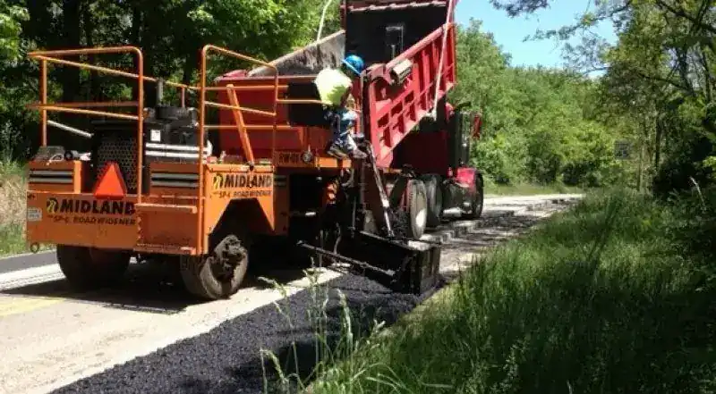 A red Shallenberger dump truck feeding a paver on a rural road, crew in hi-vis